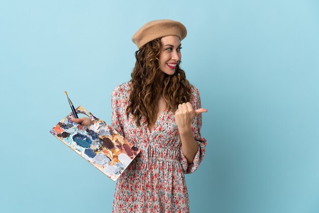 Young artist girl holding a palette isolated on blue wall pointing to the side to present a product