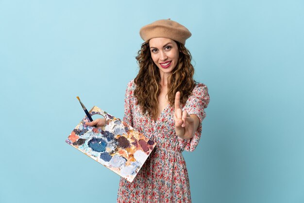 Young artist girl holding a palette isolated on blue wall showing and lifting a finger