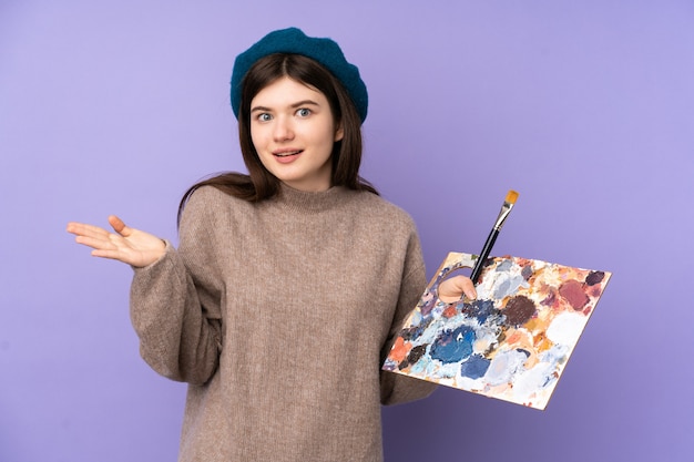  Young artist girl holding a palette over purple wall with shocked facial expression