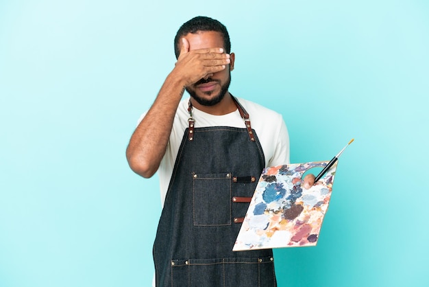 Молодой художник латиноамериканец держит палитру, изолированную на синем фоне, закрывая глаза руками. Не хочу видеть что-то