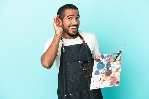 Young artist latin man holding a palette isolated on blue background listening to something by putting hand on the ear