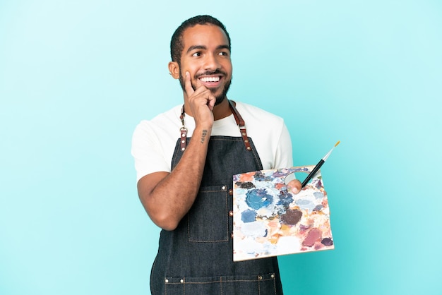 Young artist latin man holding a palette isolated on blue background thinking an idea while looking up