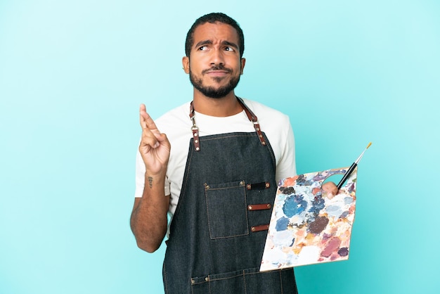 Young artist latin man holding a palette isolated on blue background with fingers crossing and wishing the best