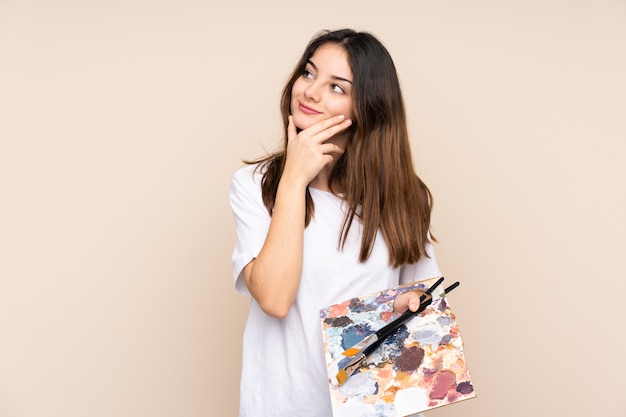 Young artist man holding a palette isolated on beige wall thinking an idea