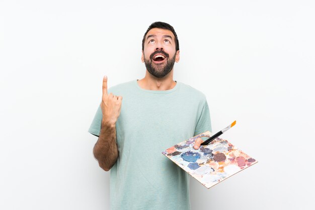 Young artist man holding a palette over isolated wall surprised and pointing up