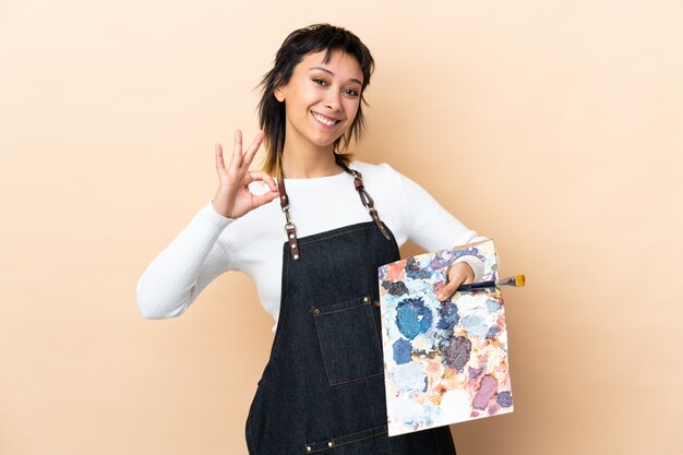 Young artist man holding a palette over wall showing an ok sign with fingers