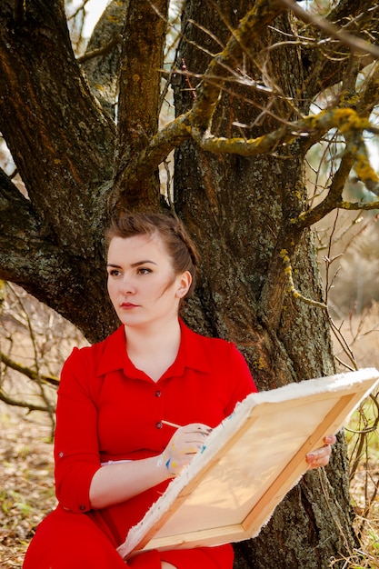 Young artist painting a landscape. Woman in red dress sitting near dry tree is drawing