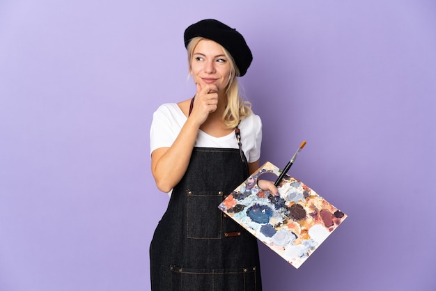Молодой художник русская женщина держит палитру, изолированную на фиолетовом фоне и смотрит вверх