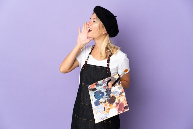 Young artist Russian woman holding a palette isolated on purple background shouting with mouth wide open to the side