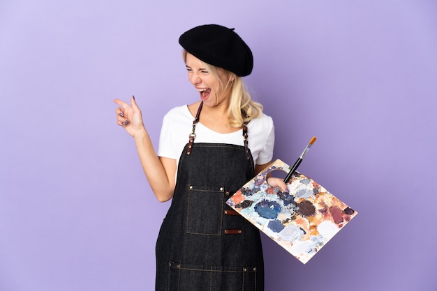 Молодая русская художница держит палитру, изолированную на фиолетовом, указывая пальцем в сторону и представляет продукт