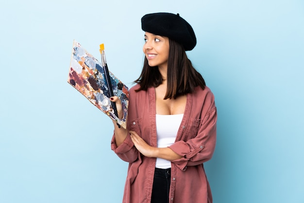 Young artist woman holding a palette over blue wall looking side