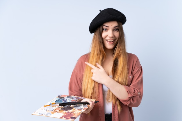 Young artist woman holding a palette on blue wall pointing finger to the side