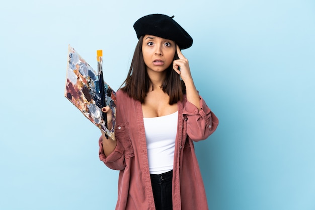 Photo young artist woman holding a palette over blue wall thinking
