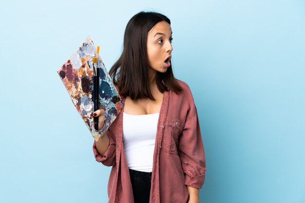 Young artist woman holding a palette over isolated blue background doing surprise gesture while looking to the side