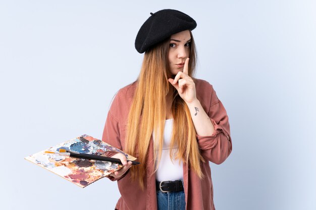 Young artist woman holding a palette isolated on blue wall doing silence gesture