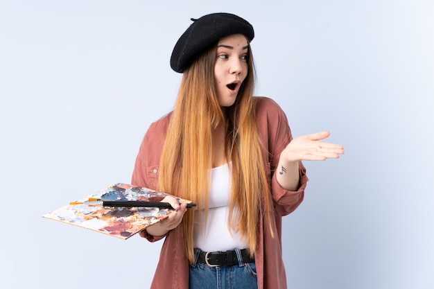 Photo young artist woman holding a palette isolated on blue wall holding copyspace imaginary on the palm