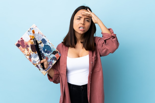 Young artist woman holding a palette over isolated blue wall looking far away with hand to look something