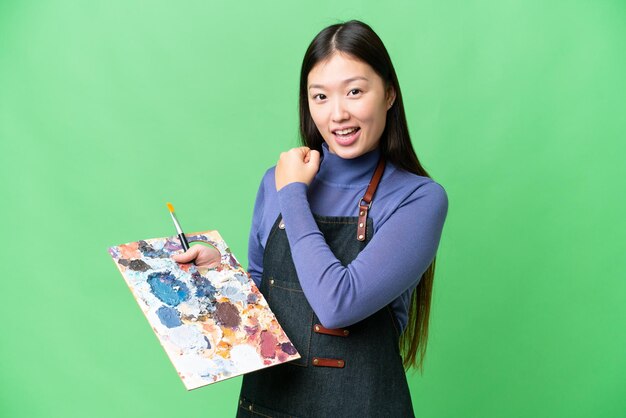 Young artist woman holding a palette over isolated chroma key background celebrating a victory