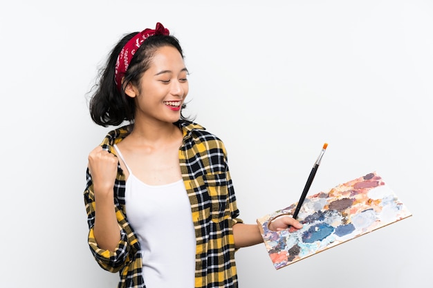 Young artist woman holding a palette over isolated white wall celebrating a victory