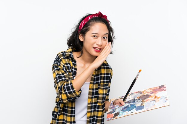 Young artist woman holding a palette over isolated white wall whispering something