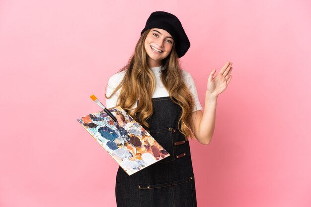 Young artist woman holding a palette on pink saluting with hand with happy expression