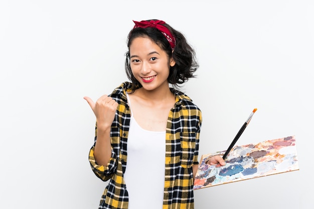 Young artist woman holding a palette over white wall pointing to the side to present a product