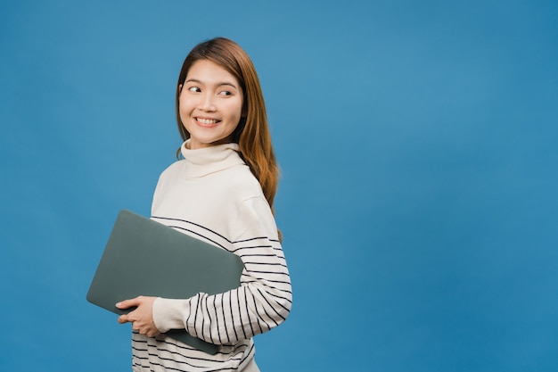 젊은 아시아 여성은 긍정적인 표정으로 노트북을 들고 활짝 웃고 행복을 느끼는 캐주얼한 옷을 입고 파란 벽에 고립되어 있습니다.