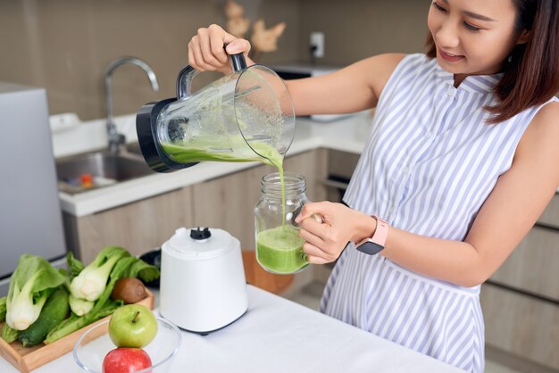 Foto giovane donna asiatica che versa frullato verde nel bicchiere nella cucina moderna