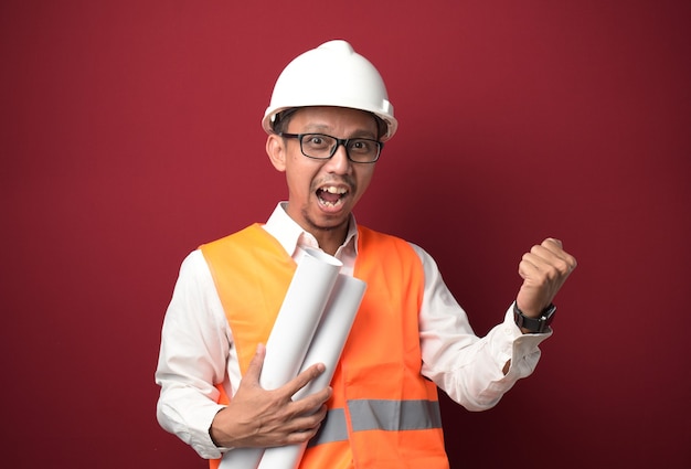 Young Asian architect wearing helmet and holding project paper plan under arm with fist clenched. 