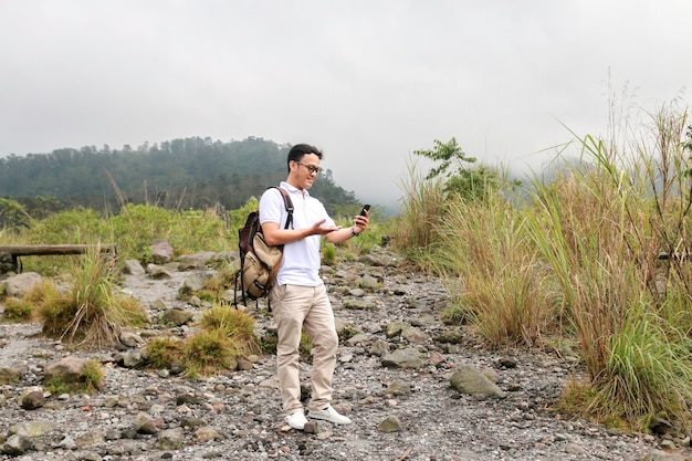若いアジアのバックパッカーの男は、山を旅行しているときにスマートフォンを見て幸せで笑顔です