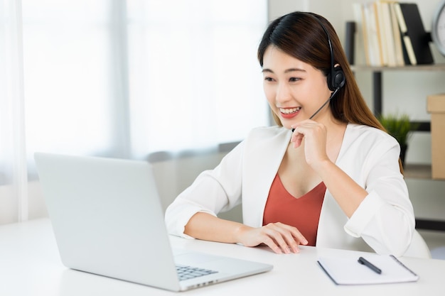 Giovane bella donna asiatica di affari che lavora con il computer portatile che si siede a casa. sorridente affascinante felice giovane femmina che fa i compiti riunione conferenza con il team a casa. indossa la cuffia guardando lo schermo