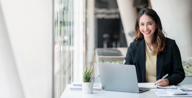 Молодая азиатская красивая и очаровательная деловая женщина улыбается и работает на ноутбуке в офисе