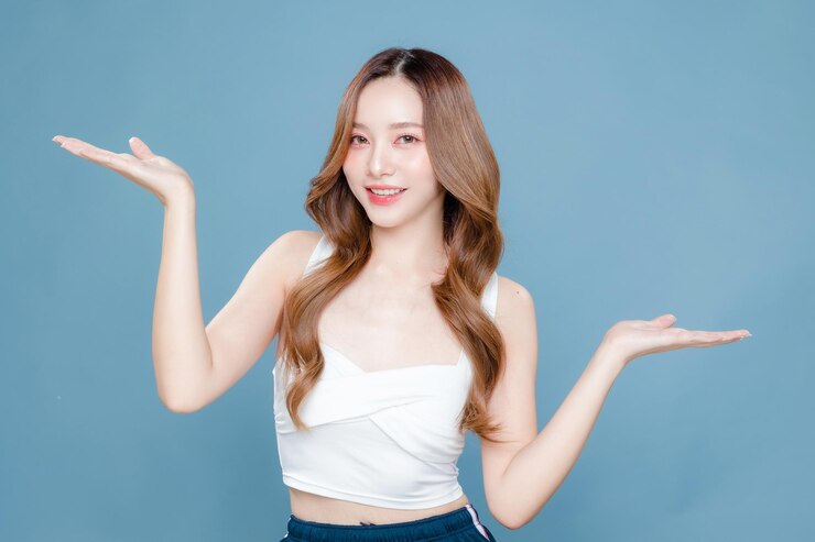 年輕的亞洲美女長髮卷曲,臉上有韓國化<unk>風格,皮膚完美清潔,在孤立的藍色背景上顯示開放的手掌,面部治療,美容整形外科