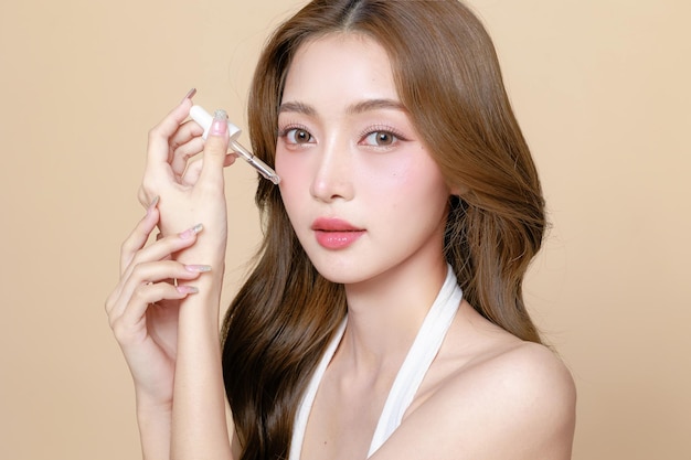 アジアの美人女性顔に韓国風のメイクアップを着用しベージュ色の背景で皮膚に血清を塗装するためにパイペットを使用しています 顔の治療美容整形外科