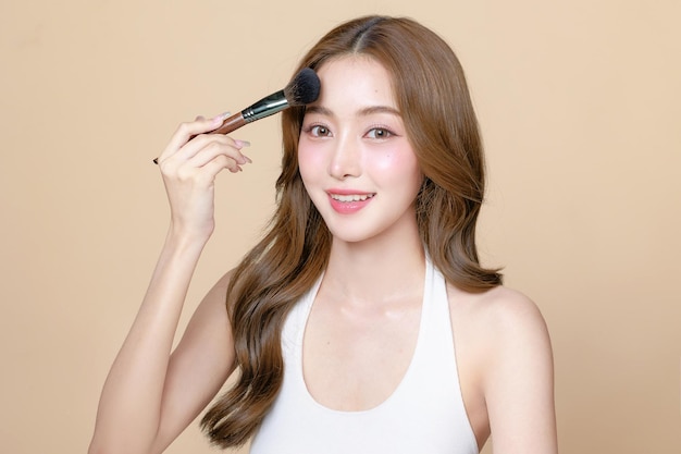 Young Asian beauty woman curly long hair with korean makeup style touch her face and perfect skin using brush on isolated beige background Facial treatment Cosmetology plastic surgery