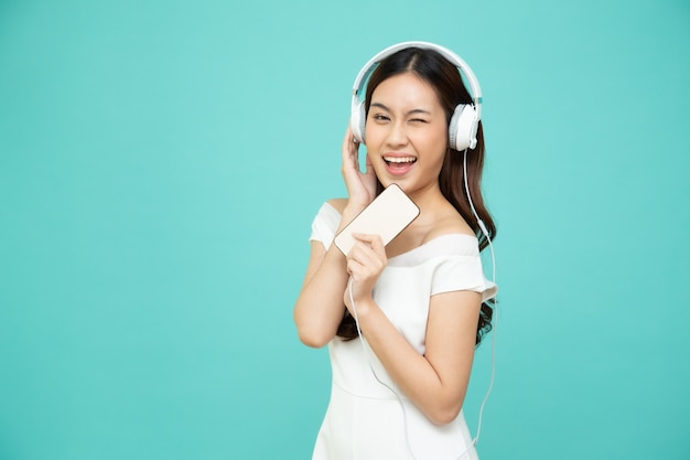 Молодая азиатская женщина красотки слушая с наушниками в приложении песни плейлиста на smartphone изолированном на зеленой стене