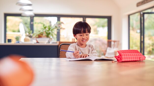 Young Asian Boy Home Schooling Working At Table In Kitchen Writing In Book