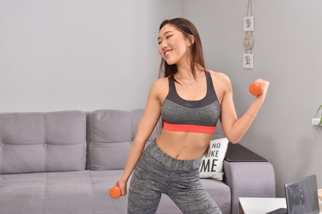 Young asian brunette woman in sportwear making home workout with two orange dumbbells.