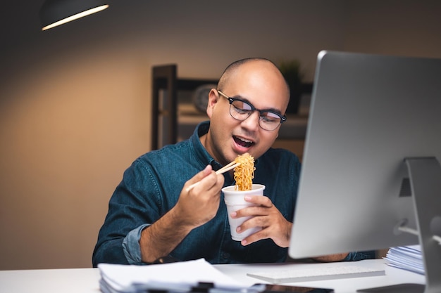 Giovane libero professionista asiatico che lavora a tarda notte ha molto fame e mangia spaghetti istantanei caldi e lavora con il computer nell'ufficio buio di notte