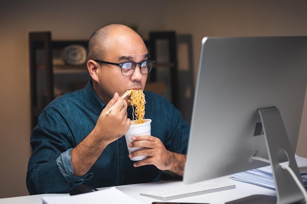 늦은 밤에 일하는 젊은 아시아 비즈니스 프리랜서 그는 매우 배고프고 뜨거운 라면을 먹고 밤에 어두운 사무실에서 컴퓨터로 일합니다