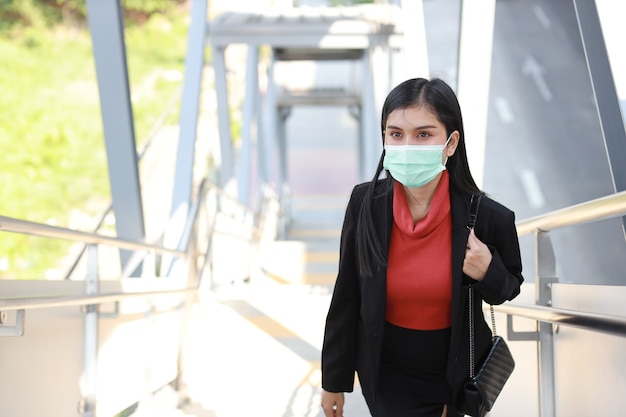 Giovane donna d'affari asiatica in abito nero da lavoro con maschera protettiva per l'assistenza sanitaria che cammina sulla passerella pubblica all'aperto. nuovo concetto di distanza normale e sociale