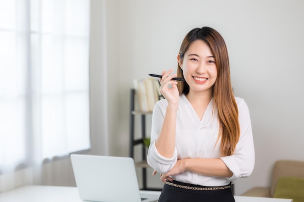 Young asian business woman standing post near desk in home office. Beautiful business female smiling sharming wearing white shirt working at home.