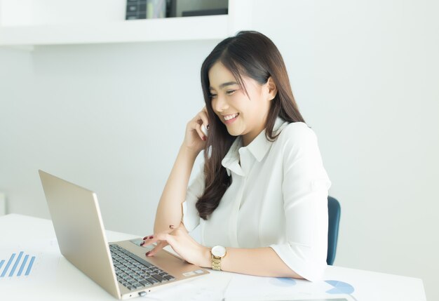 Giovane donna asiatica di affari che parla sul telefono cellulare e che sorride al suo posto di lavoro