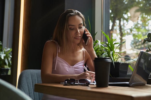 Young asian business woman writing notes in notebook at cafe working on laptop