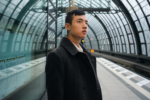 Young Asian businessman in coat thoughtfully looking away waiting train at modern subway station