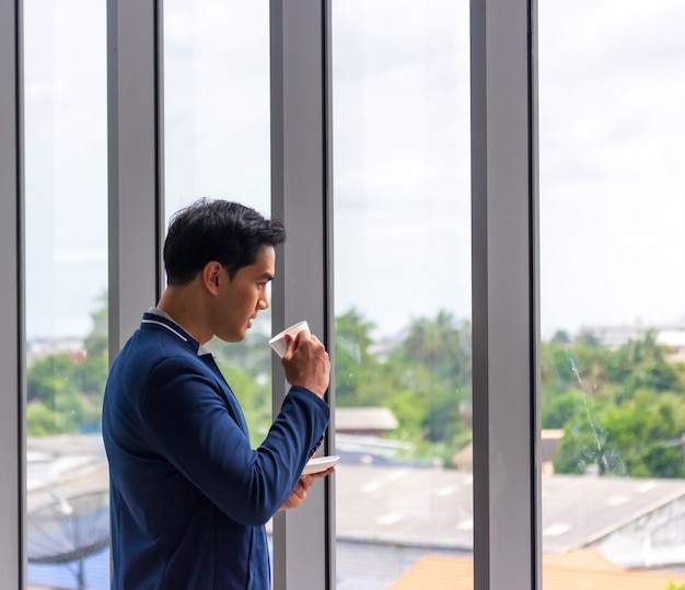 young Asian businessman drinks coffee in the office window in a comfortable