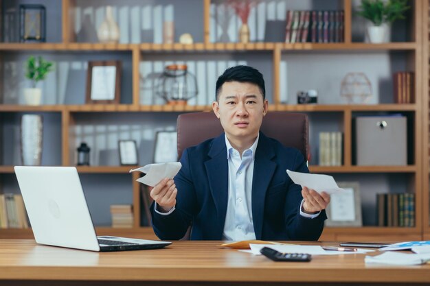 Young asian businessman man banker director sitting at desk in\
office holding bills documents embarrassed serious waving\
hands