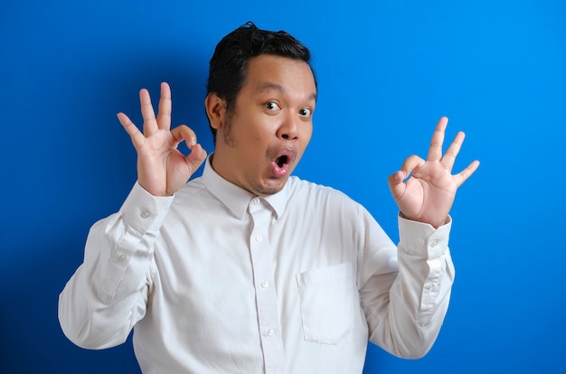 Young Asian businessman shows okay OK sign. Close up body portrait against blue wall