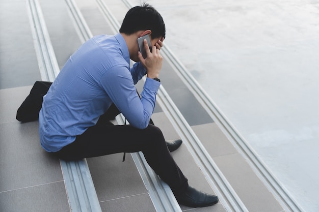 Young asian businessman sitting on stairs talking on phone are stressing.