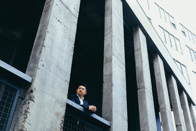 Giovane uomo d'affari asiatico in piedi vicino a un moderno edificio per uffici nero fatto di cemento, sulla terrazza del balcone con colonne